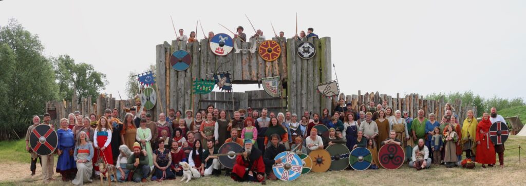 Eine größere Gruppe von Menschen in wikingerzeitlicher Kleidung hat sich vor einer hölzernen Toranlage zum Gruppenbild aufgestellt.