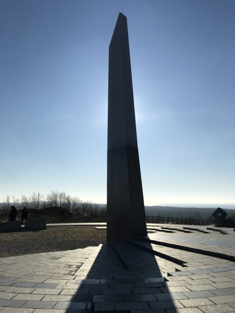 Vor dem strahlendblauen Frühjahrshimmel die dunkle Silhouette des Runenkalenders, dahinter weiter Blick über die Dünenlandschaft der Nehrung.