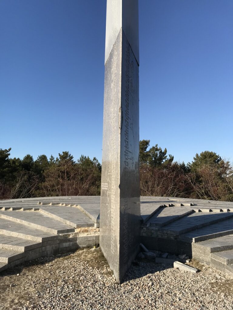Die mit Runen und Zeichen beschriftete Steinsäule von der vorderen, scharfen Kante aus fotografiert, dahinter die Kalendertreppen.