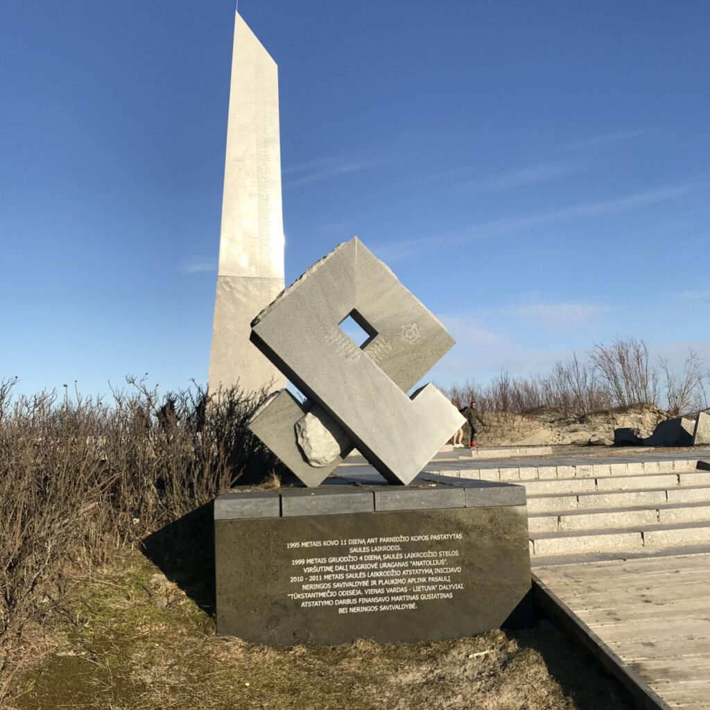 Die aufrecht stehende Säule des Runenkalender mit der Othala-Rune aus Stein im Vordergrund. Unter dem Othala ist eine kurze Beschreibung über die Anlage eingraviert.