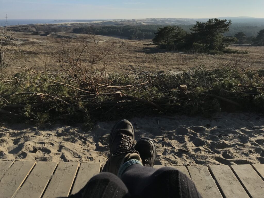 Weiter Blick über sandige Hügel, Kiefern und Meer. Der Süden der Kurischen Nehrung, im Vordergrund Beine in Wanderstiefeln.