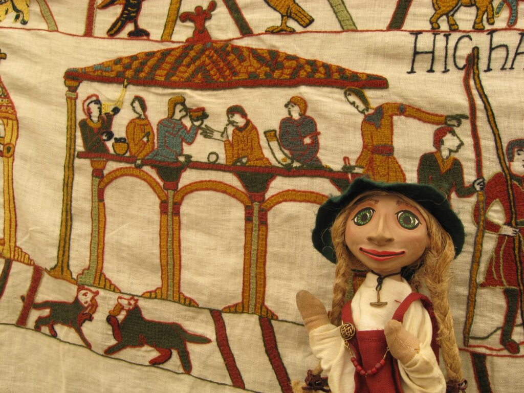 Wikinger-Handpuppe Embla neben gesticktem Detail des Bayeux-Teppichs: Darstellung eines Gebäudes mit 6 darinnen sitzenden Menschen, von denen einige Trinkhörner in Händen halten