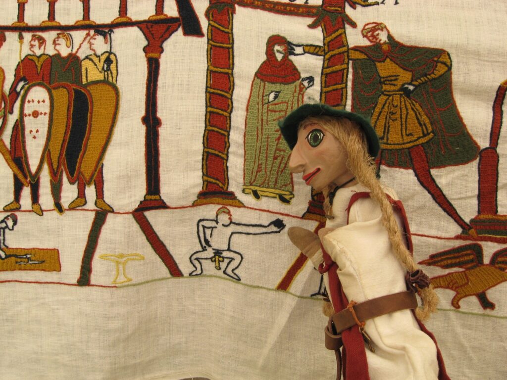 Wikinger-Handpuppe Embla vor gesticktem Detail des Bayeux-Teppichs: kleine nackte männliche Figur