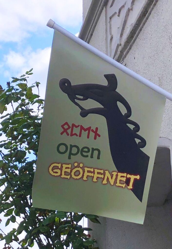 kleine Flagge an einer Hauswand, auf der ein Wikingerdrache zu sehen ist, darunter die Worte "open", einmal in Runen, und "geöffnet"