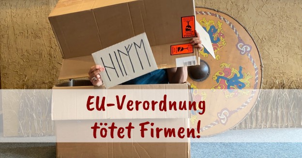 Foto zeigt zwei aufeinanderliegende Pappkartons, aus denen eine Hand hervorragt, die ein Pappschild hält. Auf diesem steht in Runen das Wort "Hilfe". Im Hintergrund ist ein bunt bemaltes Wikingerschild zu sehen.